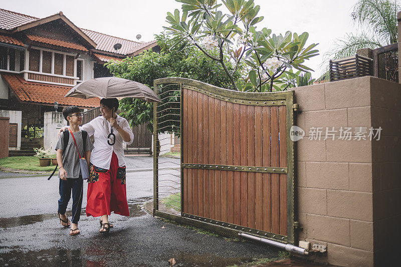 an asian chinese boy coming back from school during raining day carrying an umbrella in front of the house with his mother